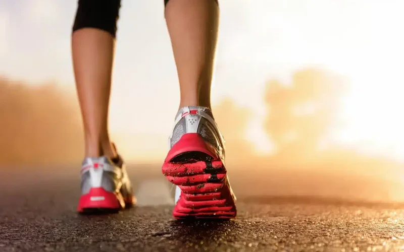 Close-up of a person’s legs wearing athletic shoes walking on a paved road at sunrise or sunset.