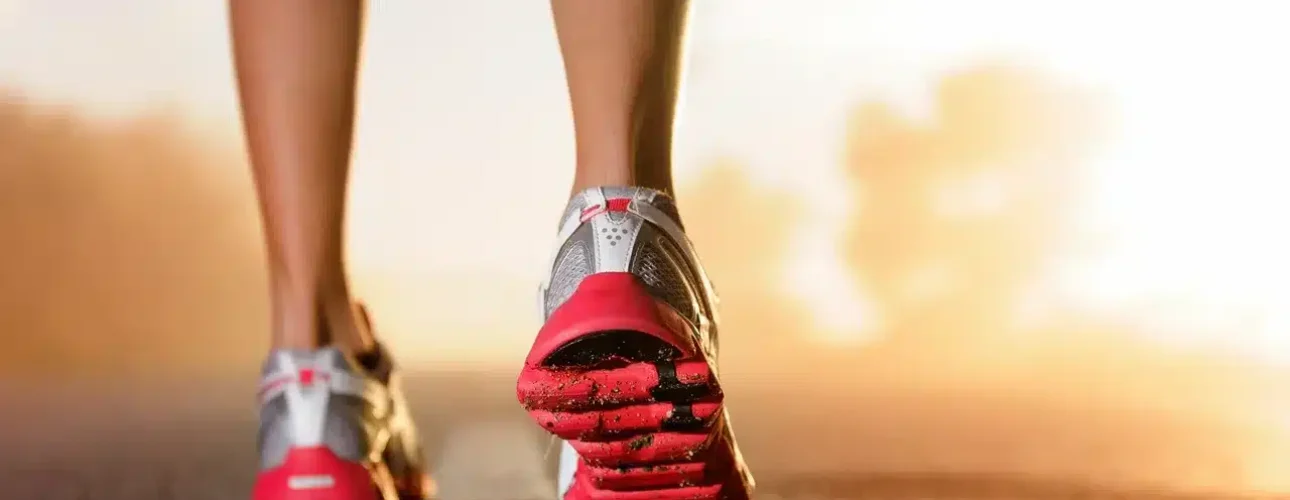 Close-up of a person’s legs wearing athletic shoes walking on a paved road at sunrise or sunset.