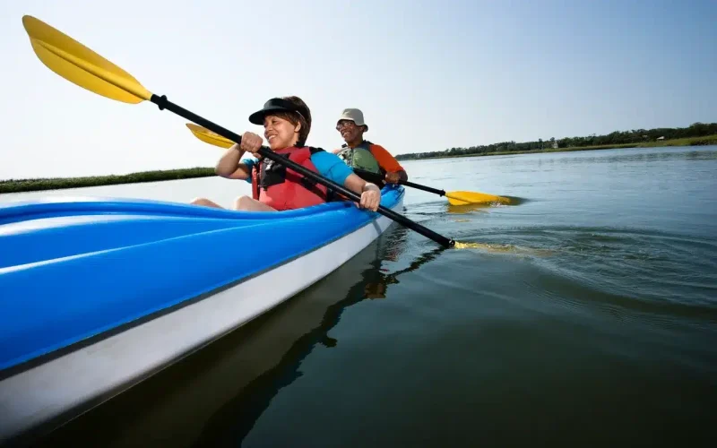 Two people wearing life jackets paddle together in a blue kayak on calm water under a clear sky.