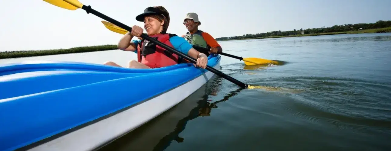 Two people wearing life jackets paddle together in a blue kayak on calm water under a clear sky.