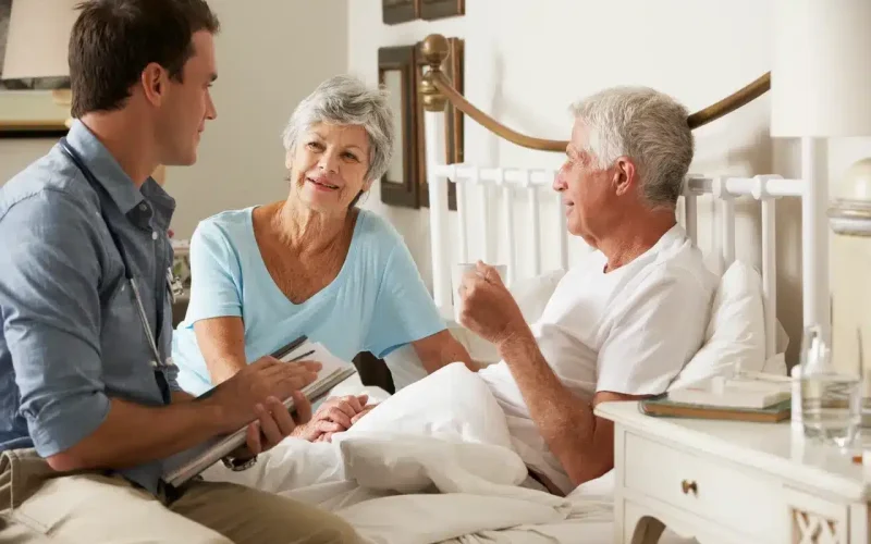 A young male healthcare professional talks to an elderly man in bed, with an elderly woman sitting beside him, in a bright, homey bedroom.