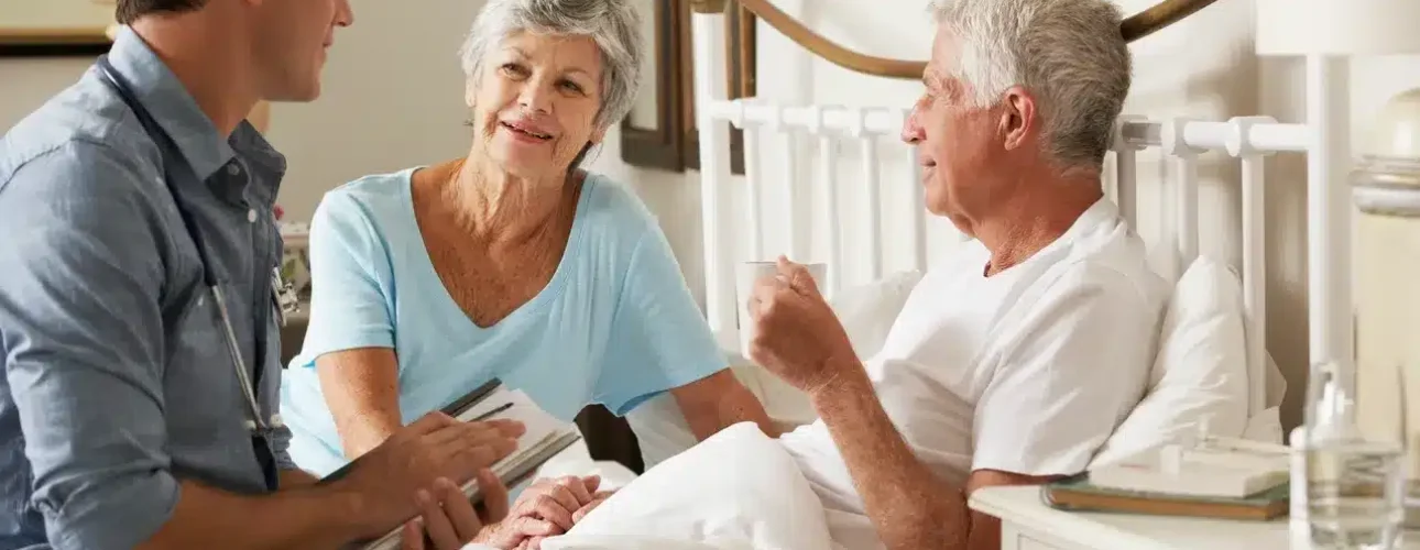 A young male healthcare professional talks to an elderly man in bed, with an elderly woman sitting beside him, in a bright, homey bedroom.