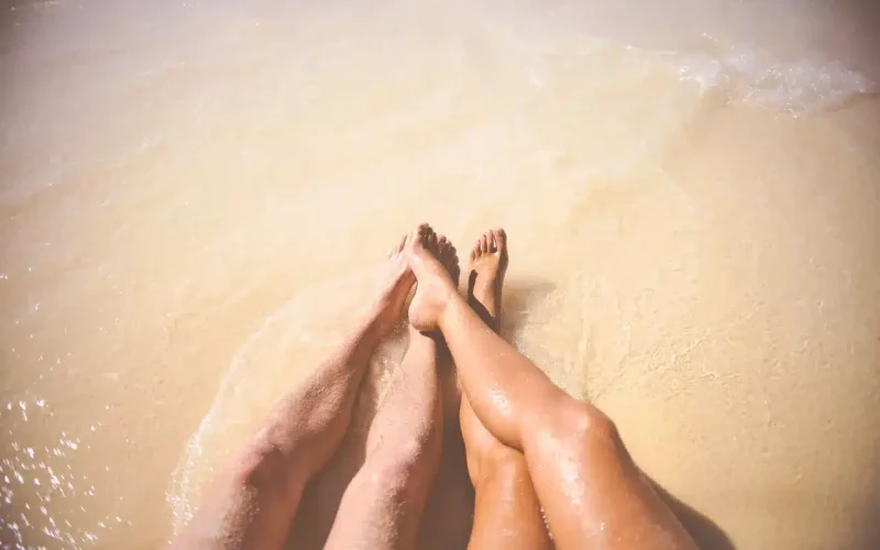 Two people sit on sandy beach with their legs extended into shallow water, where gentle waves reach their feet.