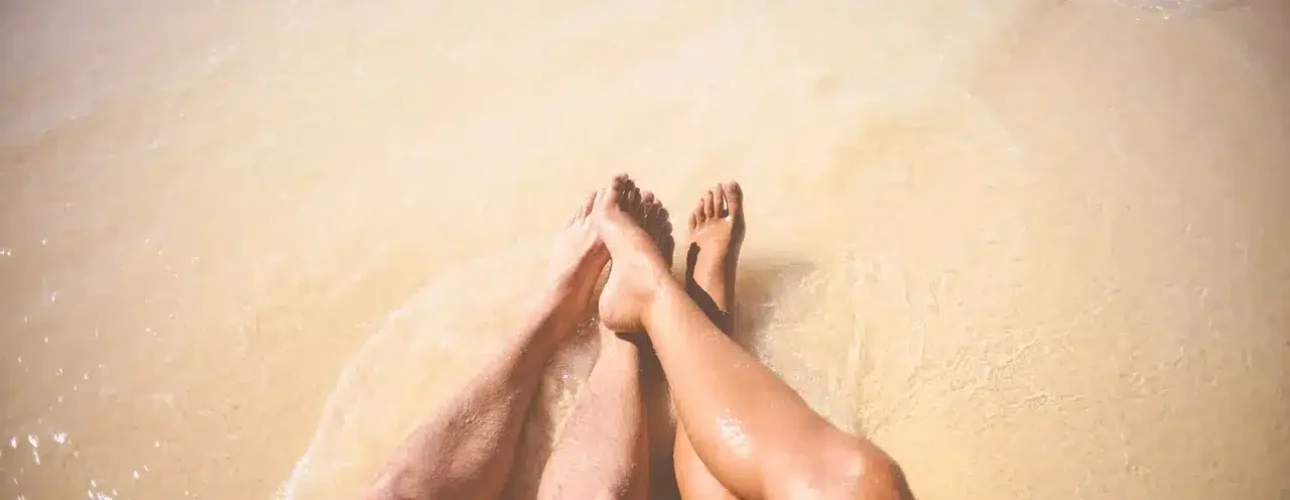 Two people sit on sandy beach with their legs extended into shallow water, where gentle waves reach their feet.
