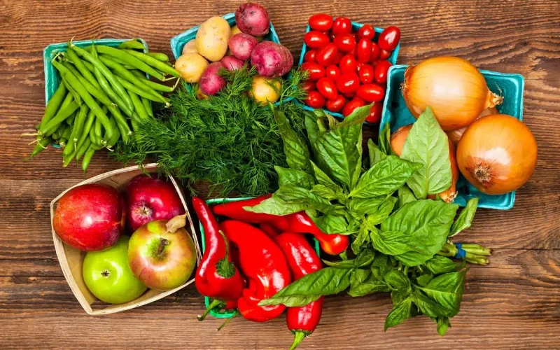 Assorted fresh vegetables and fruits, including green beans, potatoes, tomatoes, onions, apples, dill, peppers, and basil, arranged on a wooden surface.