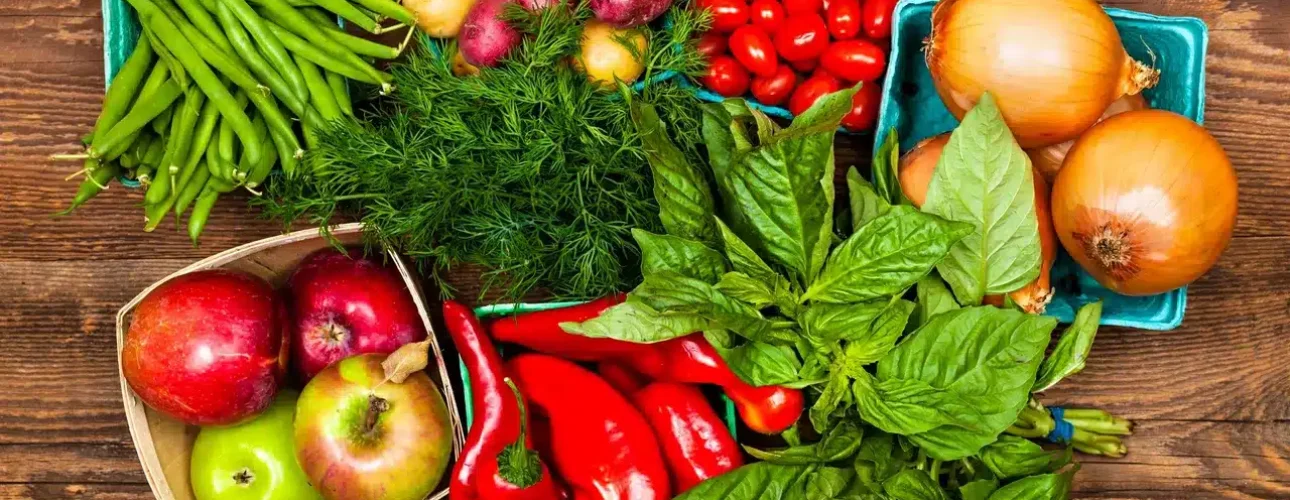 Assorted fresh vegetables and fruits, including green beans, potatoes, tomatoes, onions, apples, dill, peppers, and basil, arranged on a wooden surface.