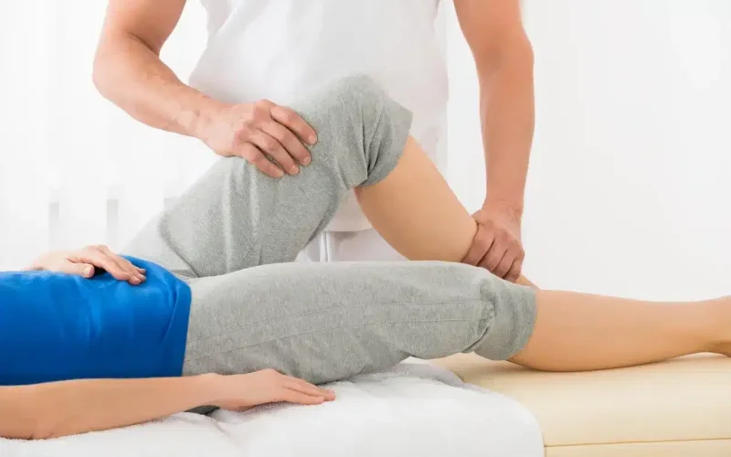 A physical therapist assists a person lying on a table by bending their knee and hip during a rehabilitation or therapy session.