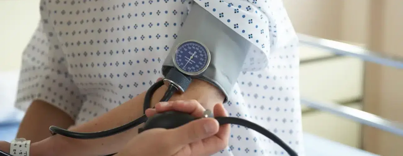 A person in a hospital gown is having their blood pressure measured with a manual sphygmomanometer by a healthcare worker.