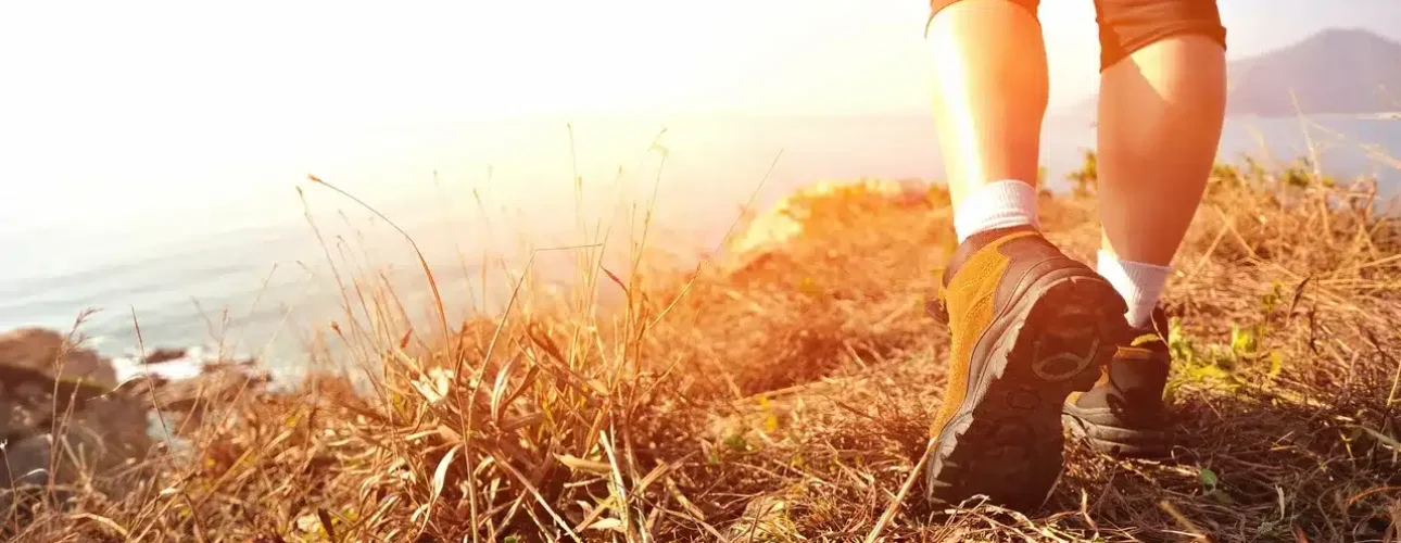 Person wearing athletic shoes and leggings walks along a dry, grassy cliffside path overlooking the ocean in bright sunlight.