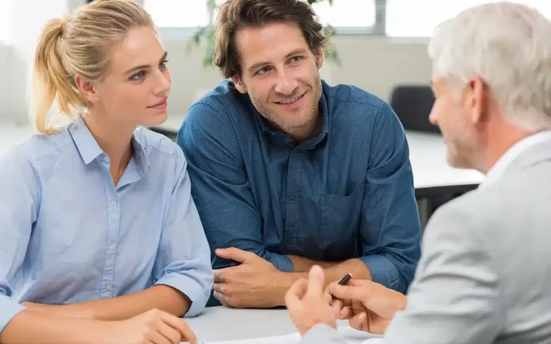 A man and woman sit across a table from an older man in a suit, engaged in a conversation in a bright office setting.