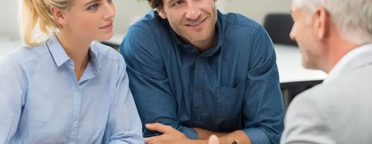 A man and woman sit across a table from an older man in a suit, engaged in a conversation in a bright office setting.