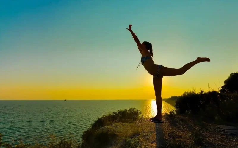 A person performs a yoga pose on one leg at a cliff's edge overlooking the sea during sunset, with arms and one leg extended.