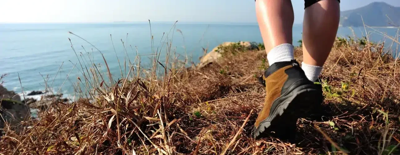 Close-up of a person wearing hiking shoes and leggings walking on dry grass near a cliff overlooking the sea on a clear day.