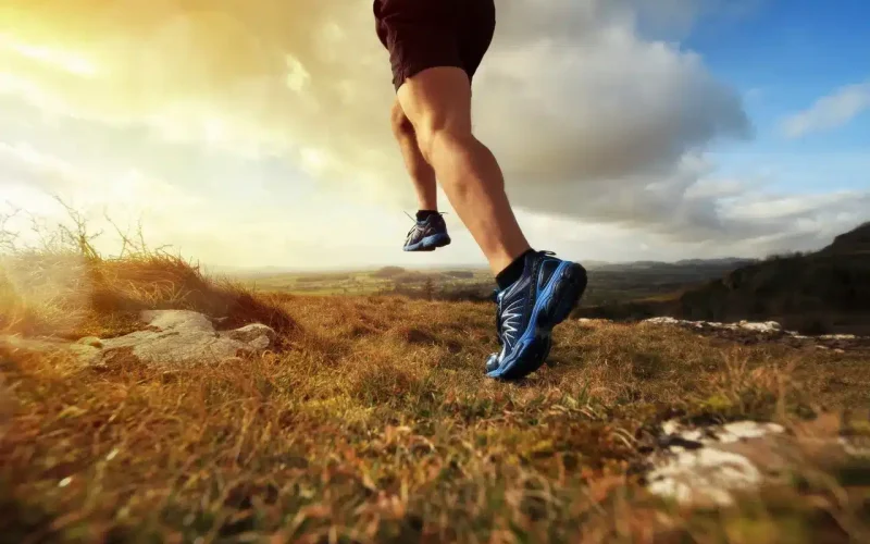 A person wearing blue running shoes jogs across a grassy field at sunset, with clouds and distant hills in the background.