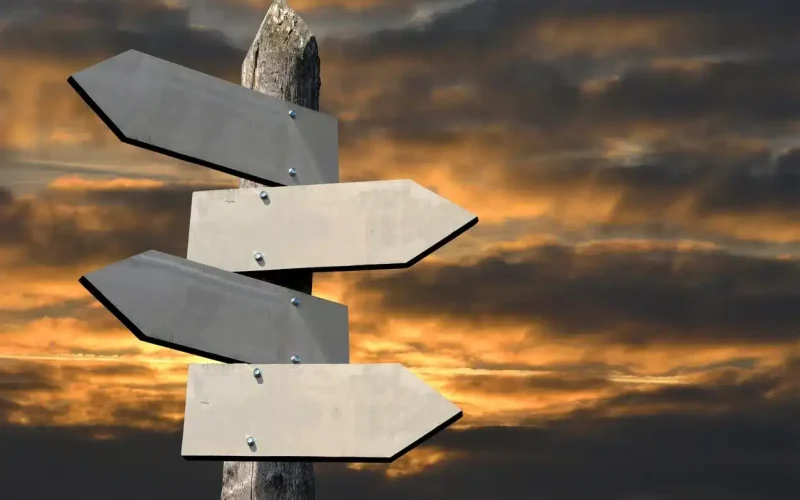 A wooden post with four blank directional signs pointing in different directions, set against a dramatic cloudy sunset sky.