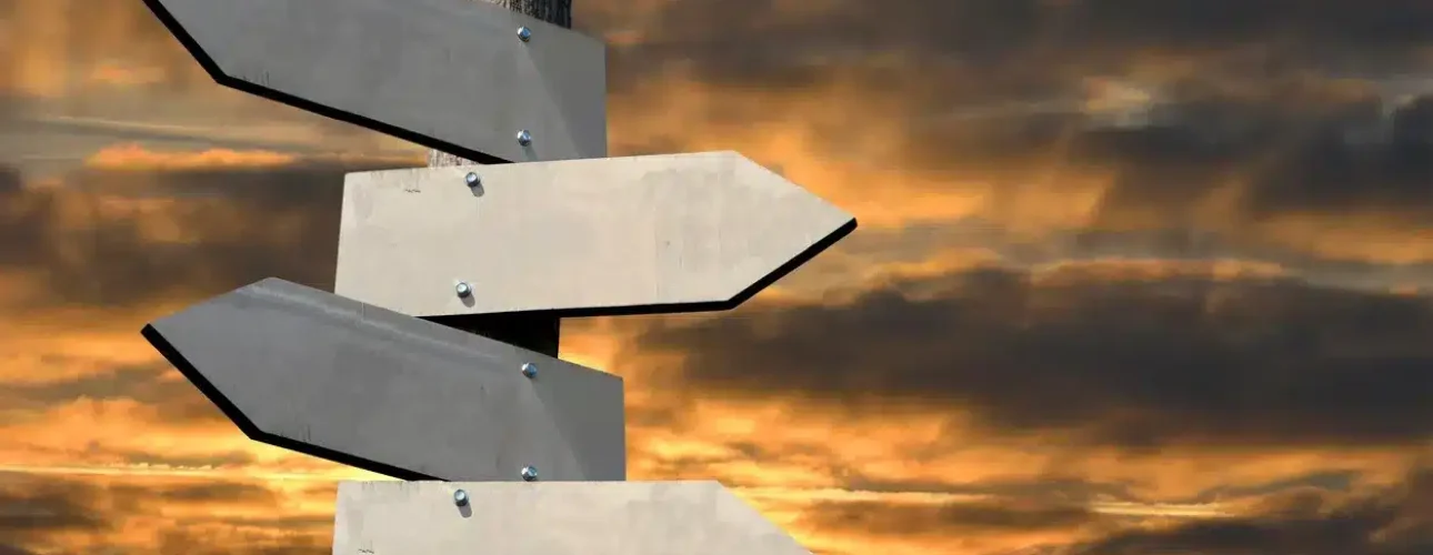 A wooden post with four blank directional signs pointing in different directions, set against a dramatic cloudy sunset sky.