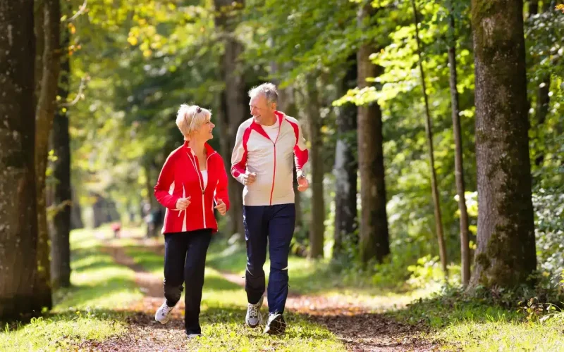 Two older adults jogging on a tree-lined path in a park, both wearing athletic clothing and looking at each other.