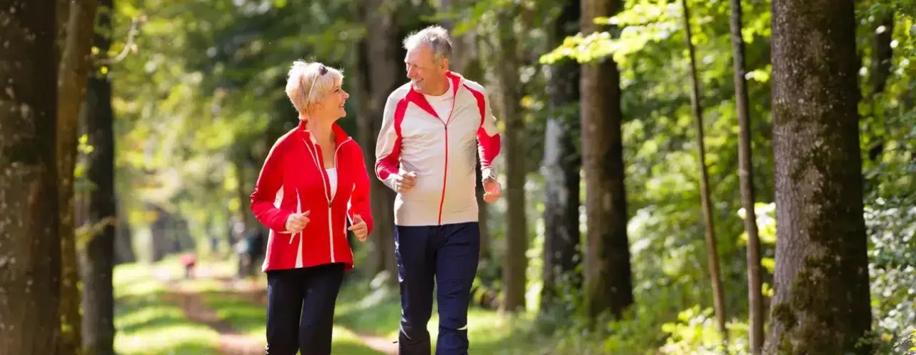 Two older adults jogging on a tree-lined path in a park, both wearing athletic clothing and looking at each other.