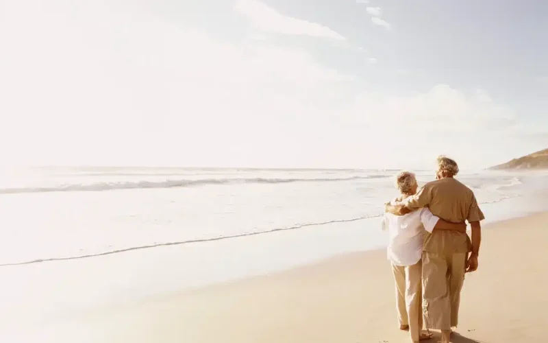 An elderly couple walks arm in arm along a sandy beach, facing the ocean under a bright, partly cloudy sky.