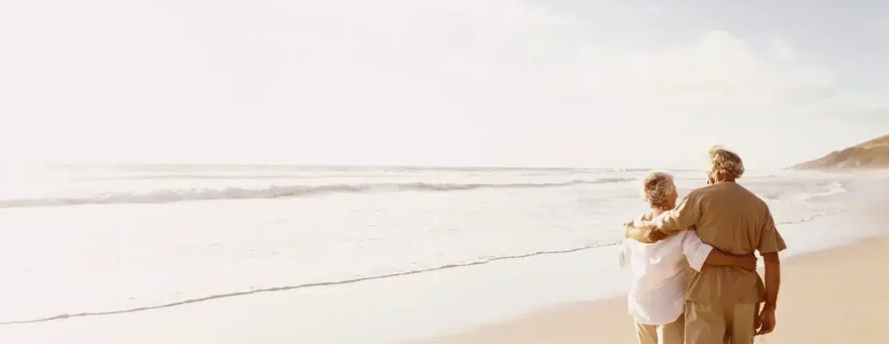 An elderly couple walks arm in arm along a sandy beach, facing the ocean under a bright, partly cloudy sky.