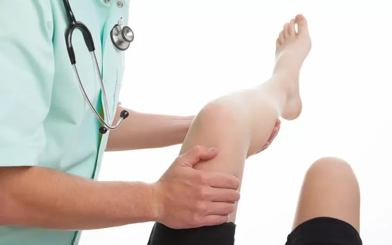 A healthcare professional examines a patient's bent knee, holding the leg with both hands.