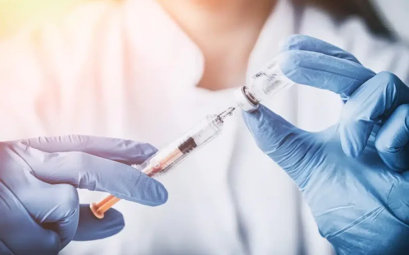 A person wearing blue gloves draws liquid from a vial into a syringe, with both hands visible and a blurred background.