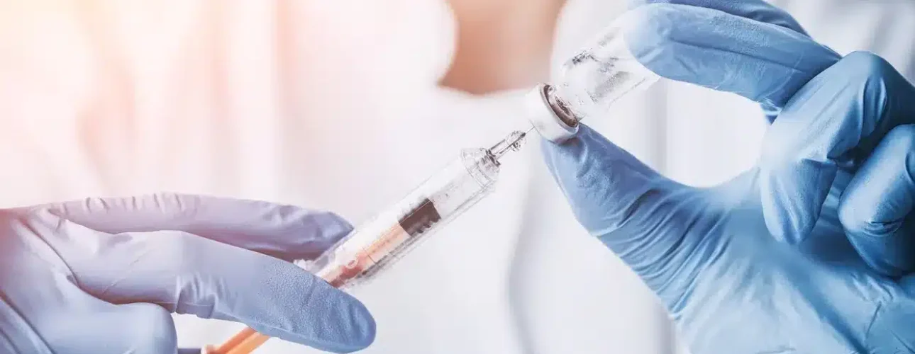 A person wearing blue gloves draws liquid from a vial into a syringe, with both hands visible and a blurred background.