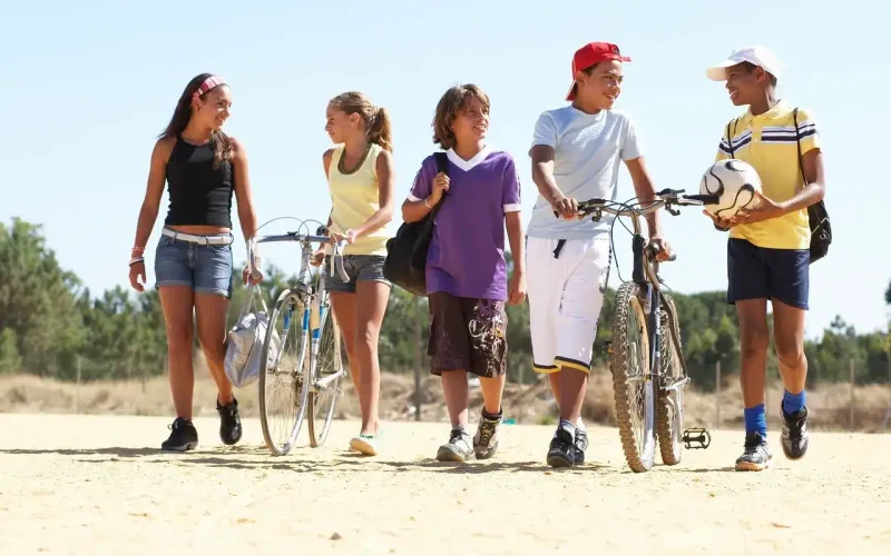 Five kids walk outdoors on a sunny day, two with bicycles and one holding a soccer ball, appearing to talk and enjoy each other's company.