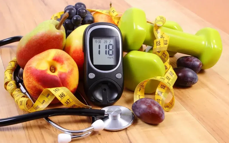 A blood glucose meter shows 118 mg/dL, surrounded by fruits, green dumbbells, a tape measure, and a stethoscope on a wooden surface.