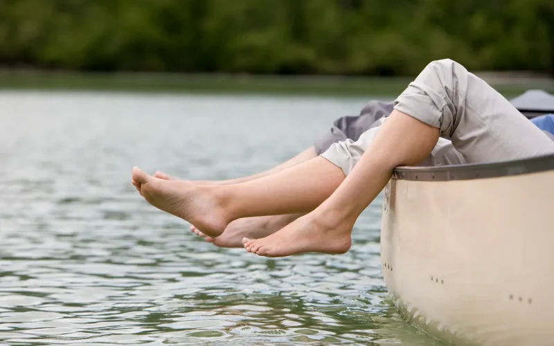 Two people sit in a canoe with their bare feet hanging over the edge, close to the water, with a blurred green shoreline in the background.