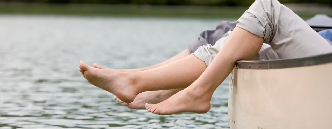 Two people sit in a canoe with their bare feet hanging over the edge, close to the water, with a blurred green shoreline in the background.