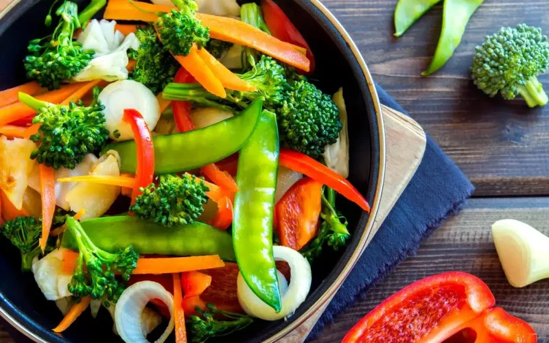 A bowl of mixed sautéed vegetables including broccoli, snow peas, red bell peppers, carrots, and onions on a wooden table.