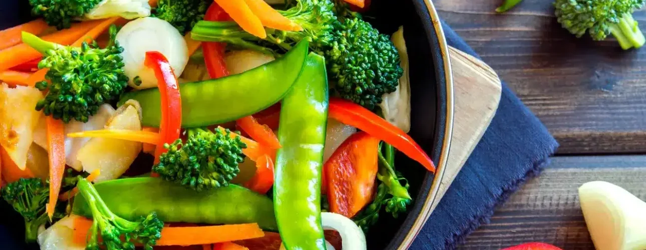 A bowl of mixed sautéed vegetables including broccoli, snow peas, red bell peppers, carrots, and onions on a wooden table.