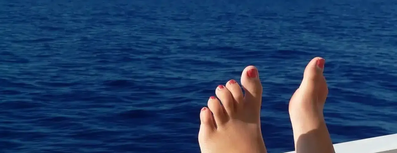 Bare feet with painted toenails rest on a white railing, overlooking a calm blue sea under a clear sky.