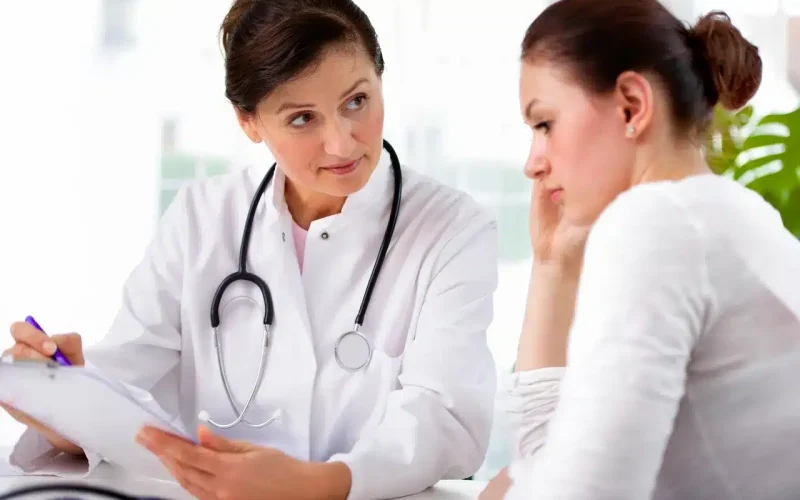 A doctor wearing a stethoscope consults with a worried patient, listening attentively and taking notes on a clipboard.