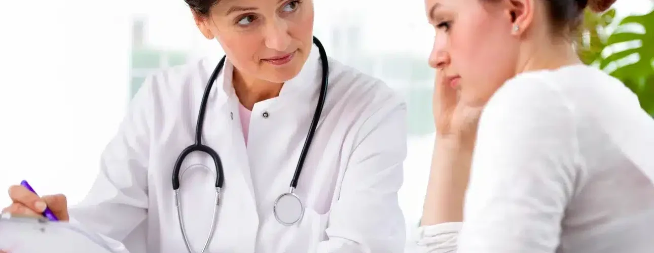 A doctor wearing a stethoscope consults with a worried patient, listening attentively and taking notes on a clipboard.