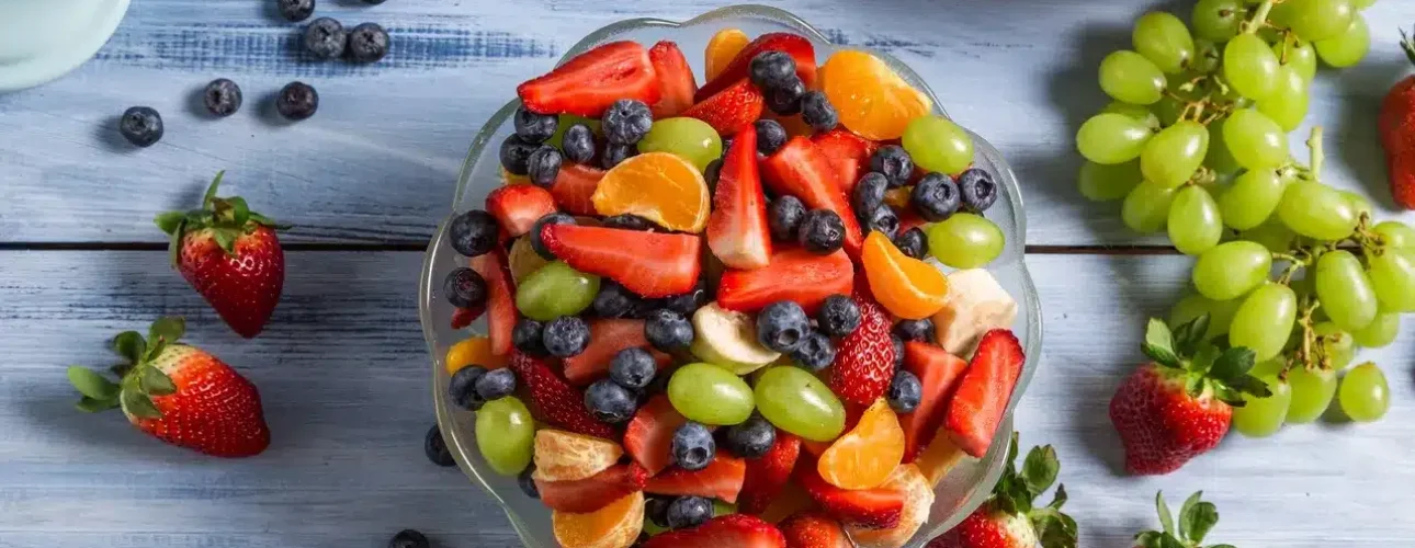 A glass bowl filled with sliced strawberries, blueberries, green grapes, and orange segments sits on a blue wooden table with more fruit scattered around.