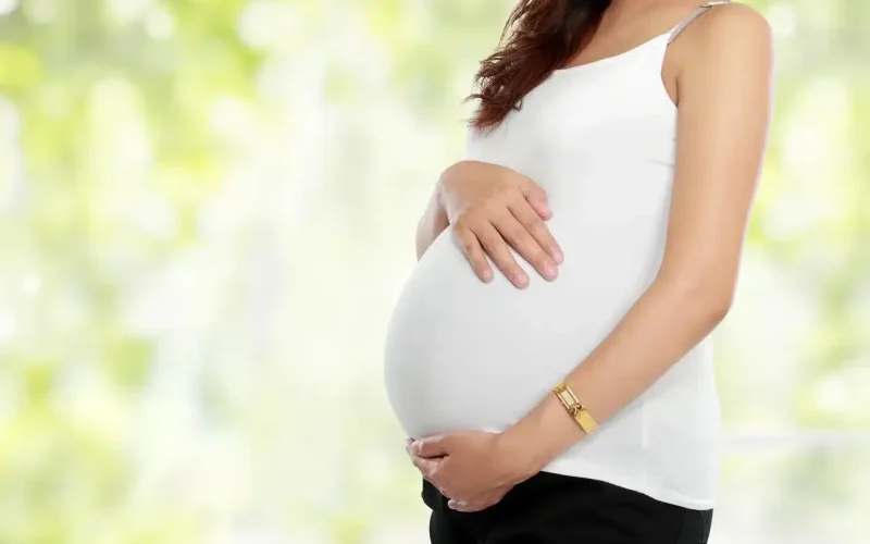 A pregnant woman in a white sleeveless top and black pants gently cradles her belly with both hands, standing against a blurred green background.