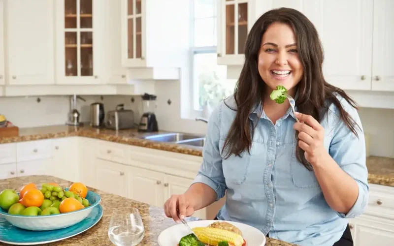 Woman sitting at a kitchen table, smiling and holding a fork with broccoli, with a plate of vegetables and a glass of water in front of her.