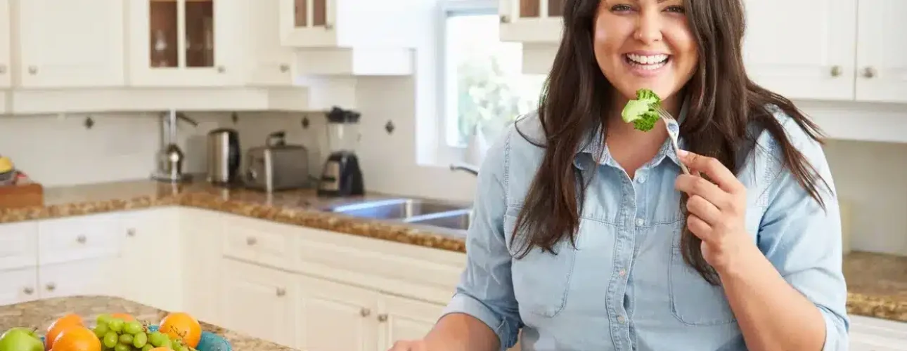 Woman sitting at a kitchen table, smiling and holding a fork with broccoli, with a plate of vegetables and a glass of water in front of her.