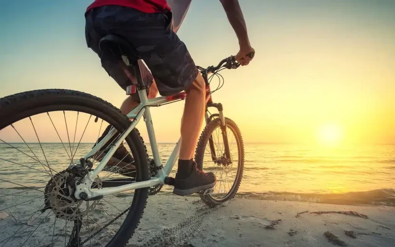 Person riding a bicycle on a sandy beach near the ocean, facing a bright sunset with clear skies.