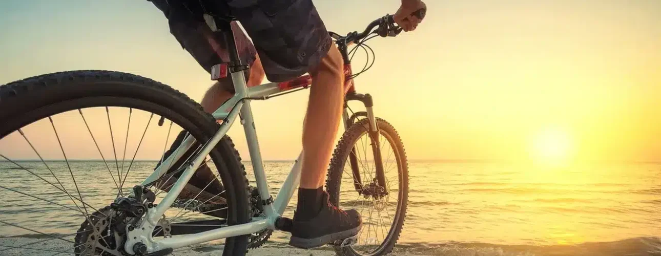 Person riding a bicycle on a sandy beach near the ocean, facing a bright sunset with clear skies.