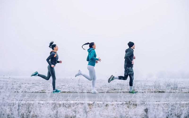Three people jog on a paved path through a frosty, foggy landscape, wearing athletic clothing suitable for cold weather.
