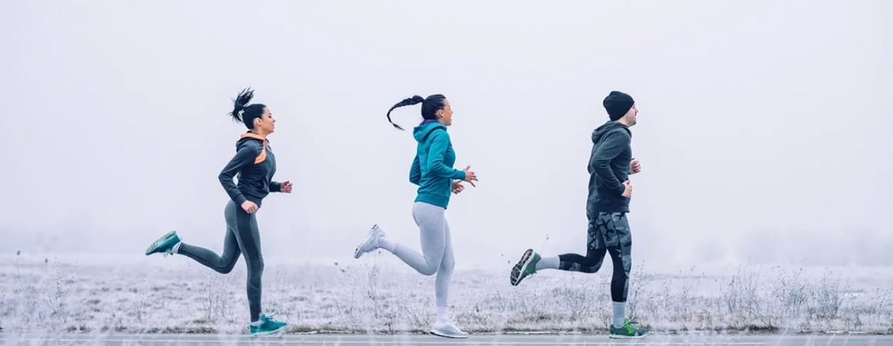 Three people jog on a paved path through a frosty, foggy landscape, wearing athletic clothing suitable for cold weather.