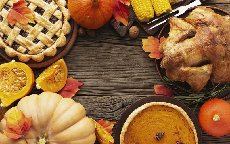 Thanksgiving meal with roast turkey, pumpkin pie, lattice pie, corn on the cob, pumpkins, and autumn leaves on a wooden table.
