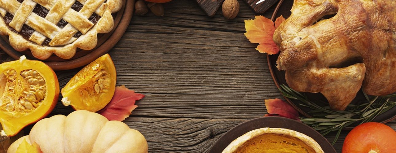 Thanksgiving meal with roast turkey, pumpkin pie, lattice pie, corn on the cob, pumpkins, and autumn leaves on a wooden table.