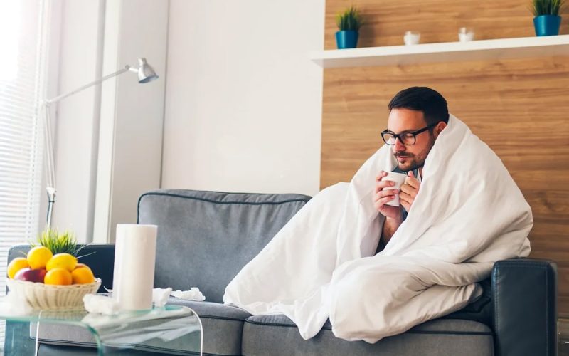 A man sits on a gray sofa, wrapped in a white blanket, holding a mug. A bowl of fruit and a tissue box are on the table in front of him in a modern living room.