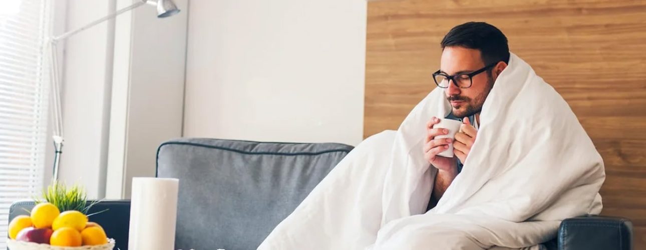A man sits on a gray sofa, wrapped in a white blanket, holding a mug. A bowl of fruit and a tissue box are on the table in front of him in a modern living room.