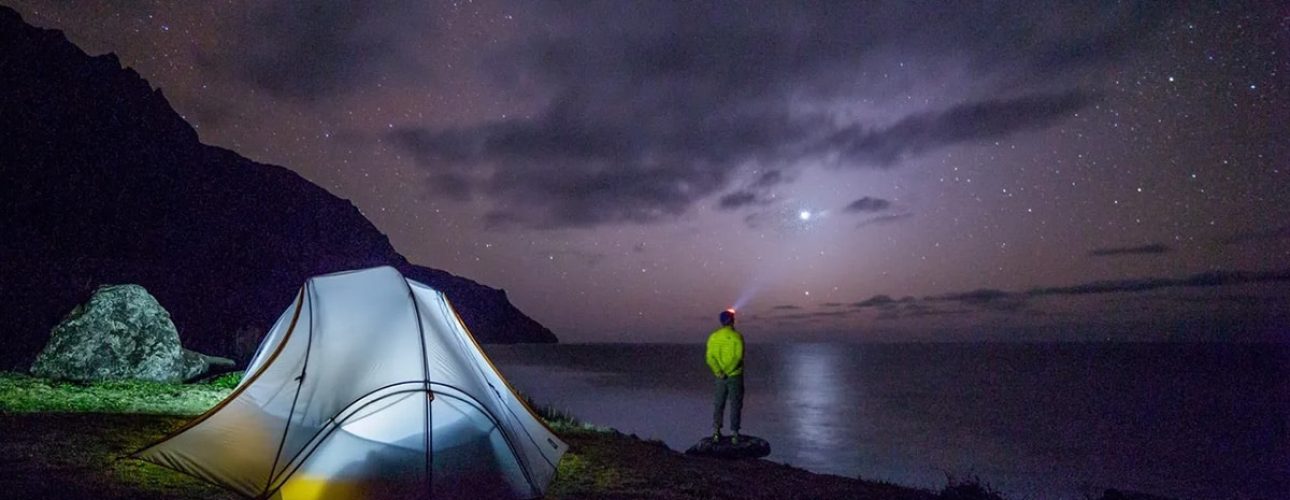 A person stands near a glowing tent on a cliff overlooking the ocean at night, with stars and clouds visible in the sky.