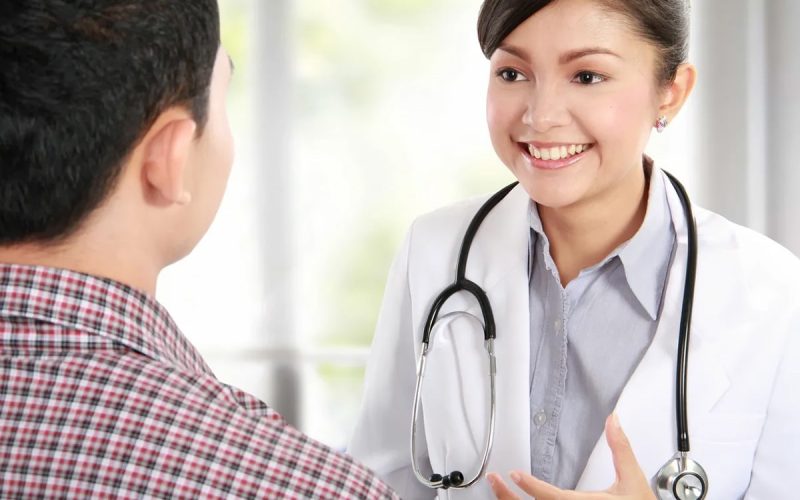 A doctor with a stethoscope smiles and speaks to a patient wearing a plaid shirt during a medical consultation.
